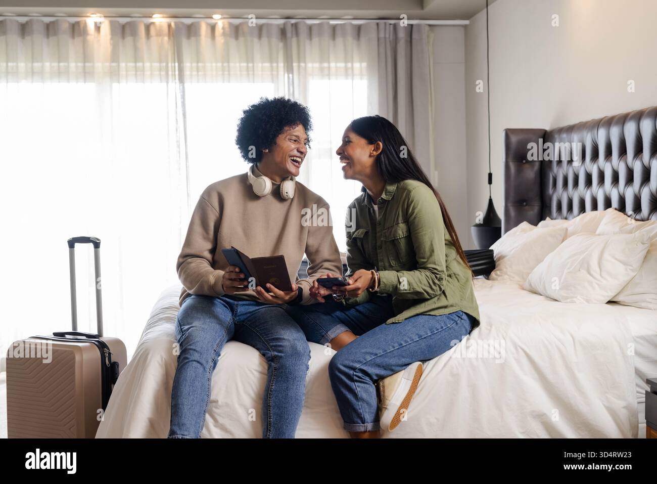 Rire couple diversifié assis sur un lit soigneusement fait dans la chambre d'hôtel, avec valise et passeports Banque D'Images