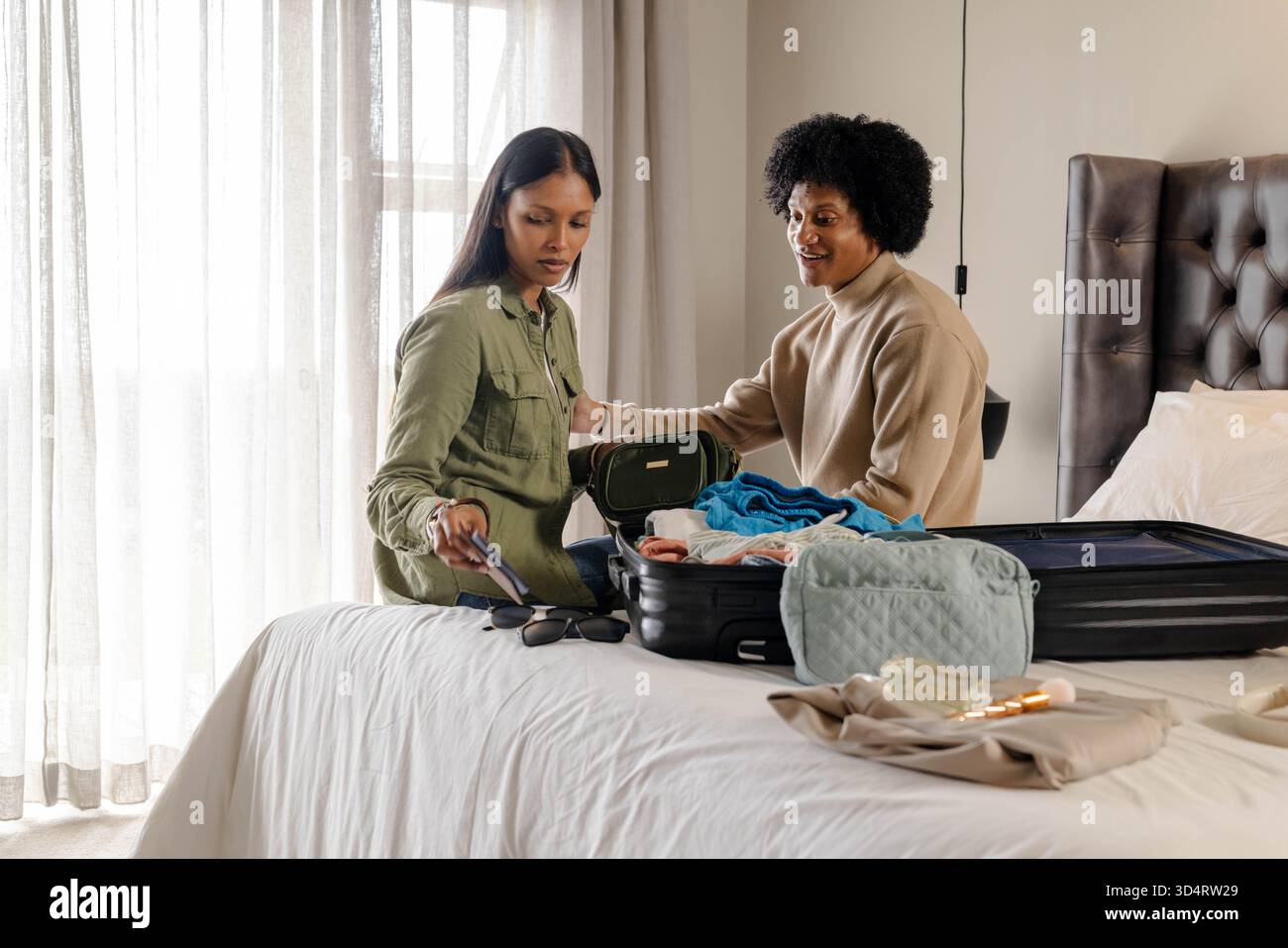 Emballage de couple diversifié assis sur le lit dans une chambre à coucher légèrement éclairée, avec valise noire rigide Banque D'Images