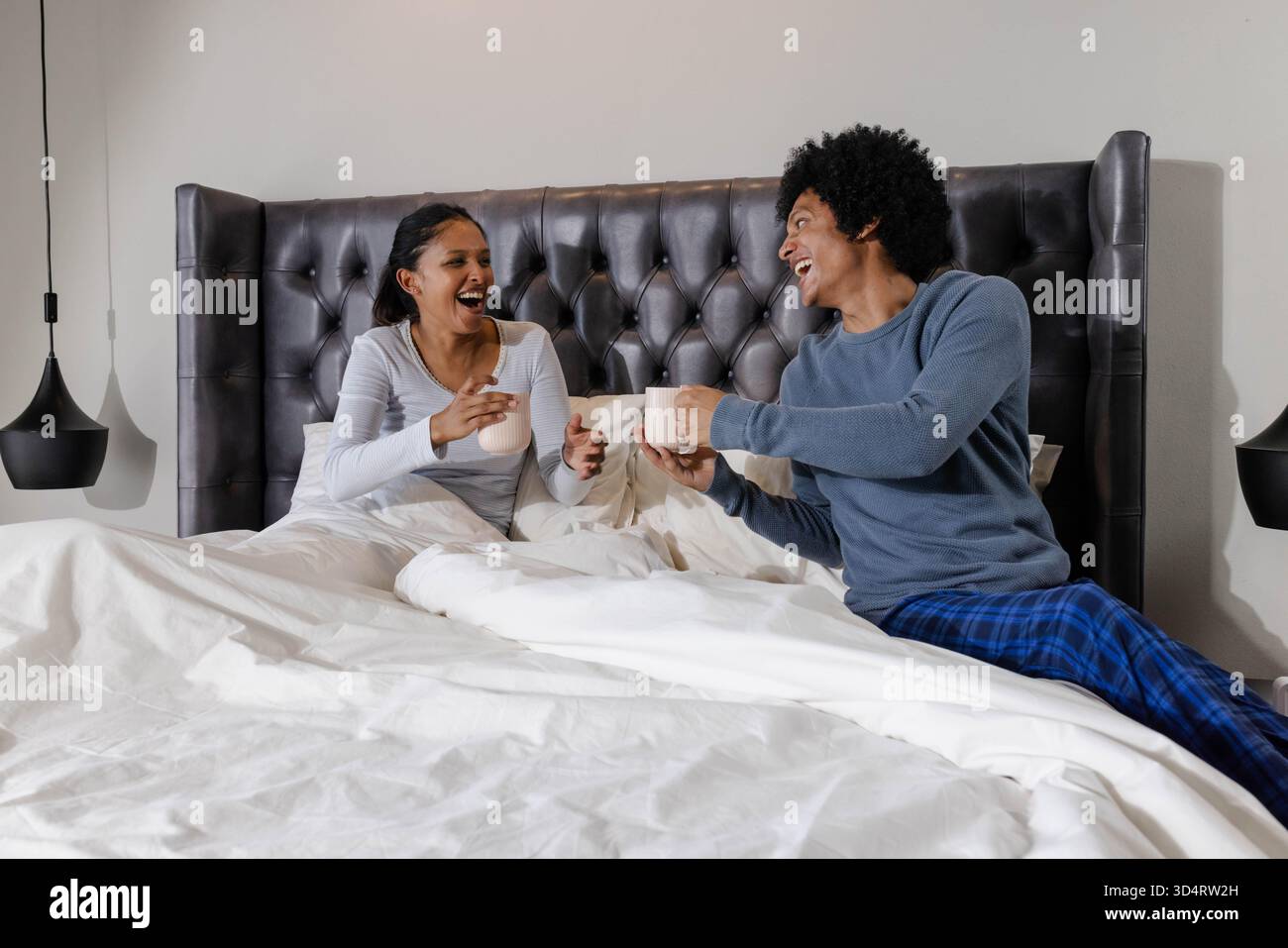 Tasses de rires de couple variées et rire dans le lit non fait dans la chambre avec tête de lit et lampes suspendues Banque D'Images