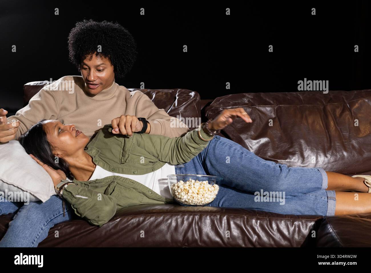 Relaxant couple diversifié câlin sur canapé en cuir sur fond noir, avec bol en verre de pop-corn Banque D'Images