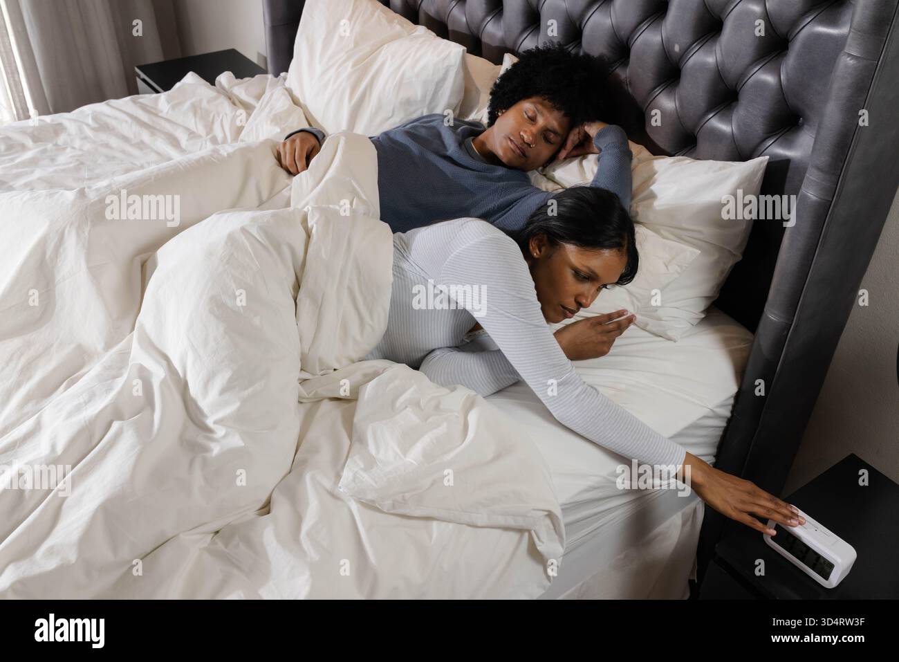 Couché couple diversifié se reposant sous couette blanche dans une chambre moderne, atteignant pour réveil Banque D'Images