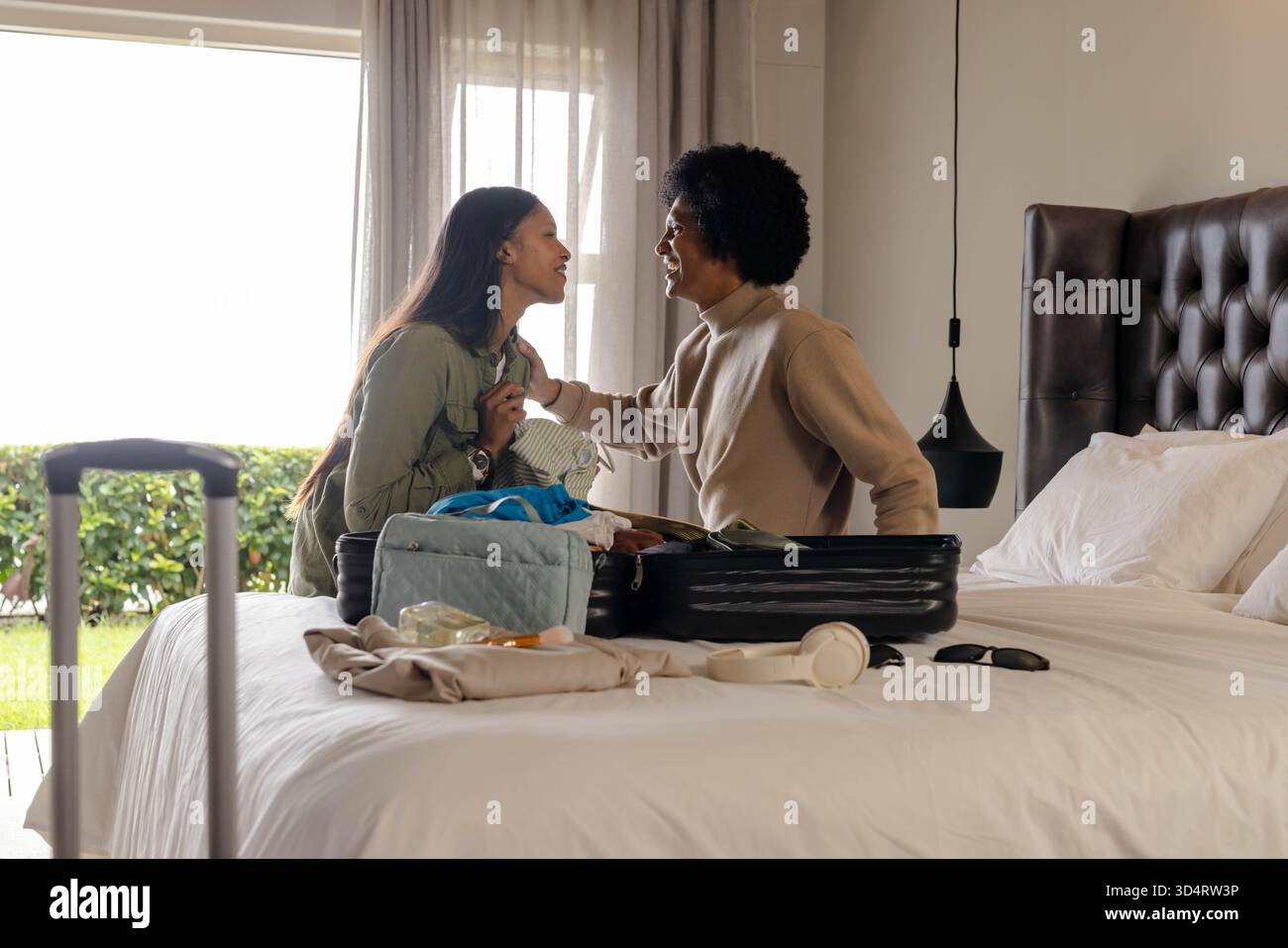 Tri divers amis féminins emballant la valise dans une chambre lumineuse, avec des écouteurs et des lunettes de soleil Banque D'Images
