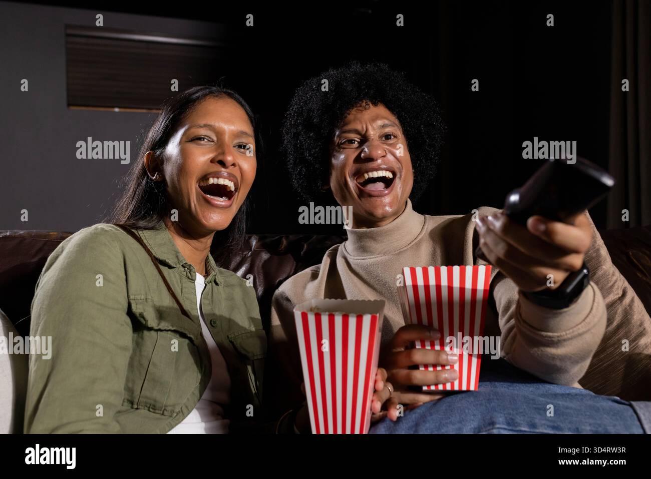 Rire divers amis assis sur le canapé en cuir brun dans le cinéma maison, avec pop-corn et télécommande Banque D'Images