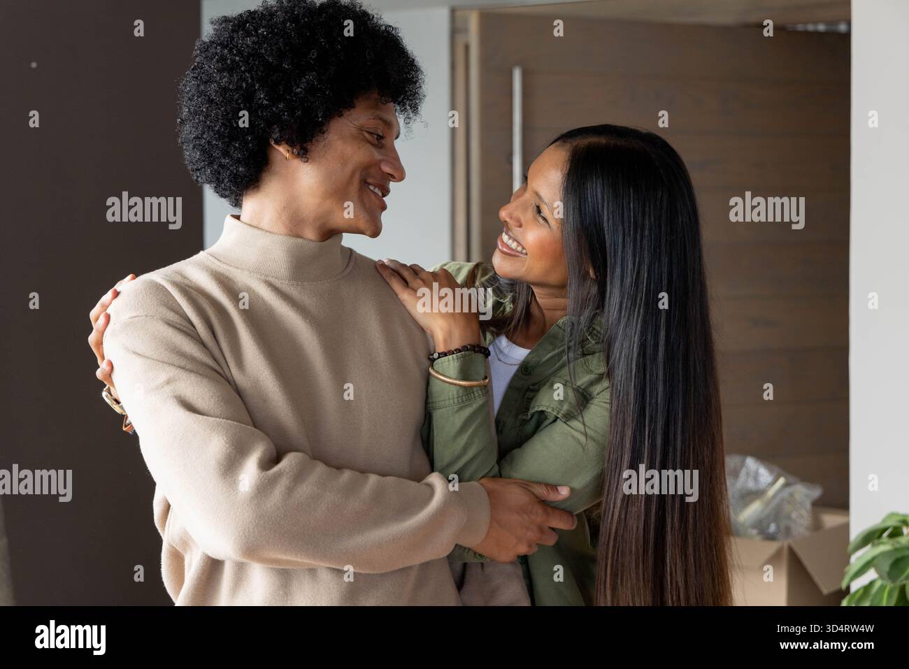 Embrasser un couple diversifié debout près de la porte d'entrée en bois à la maison, avec boîte en carton et plante Banque D'Images