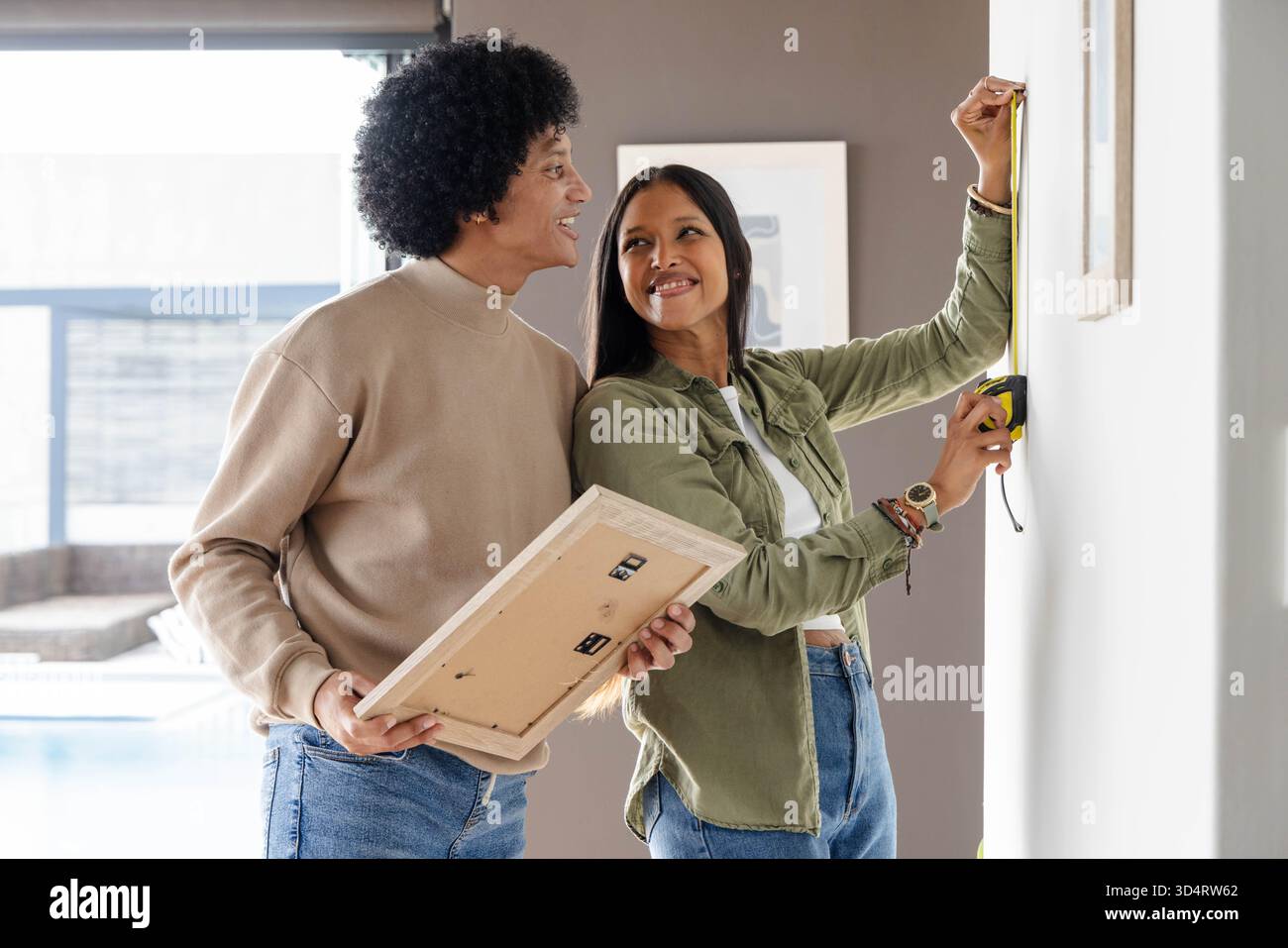 À l'aide d'un ruban à mesurer jaune, un couple diversifié accrochant l'image encadrée sur le mur neutre dans le salon Banque D'Images