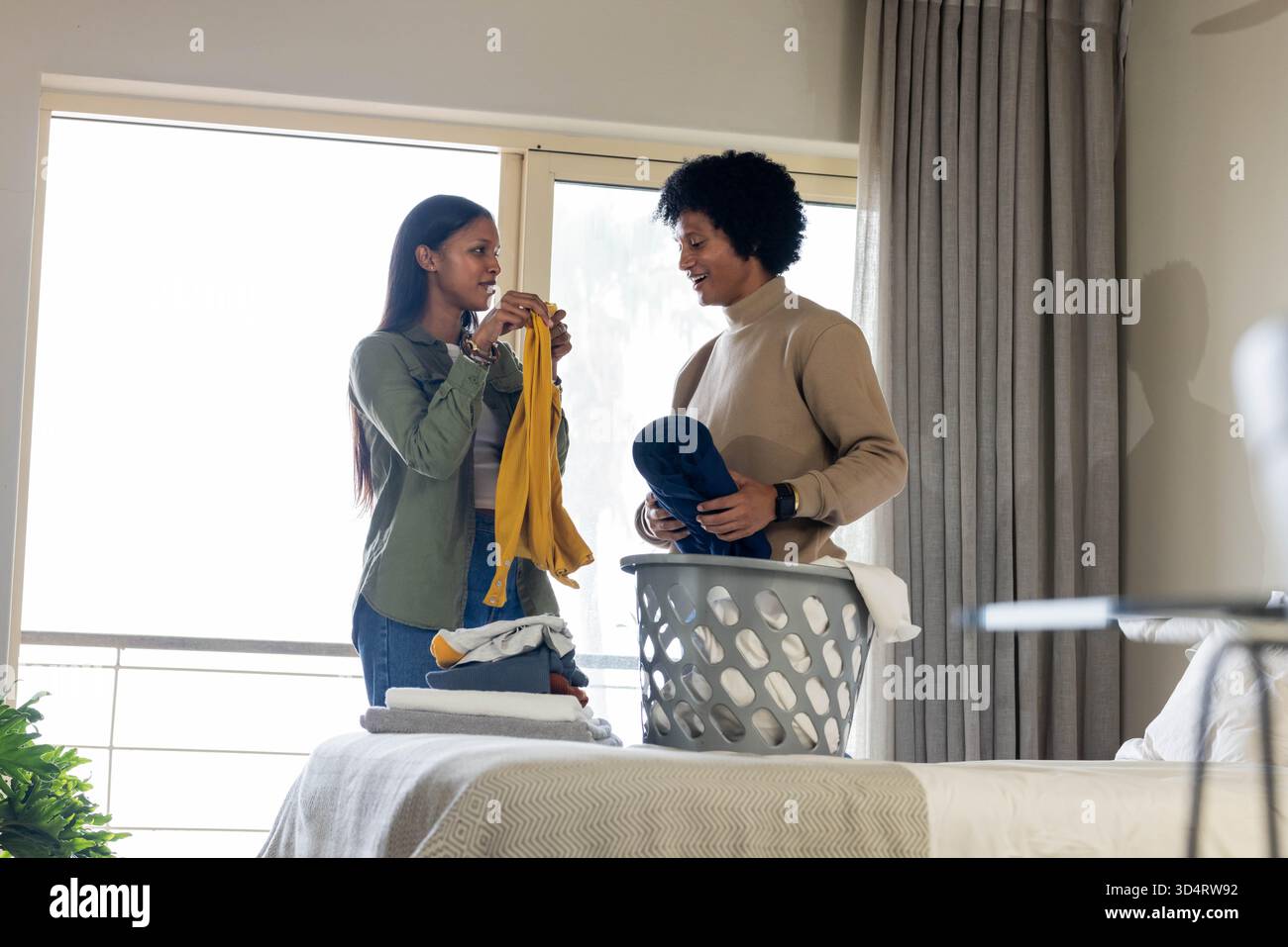 Linge pliant divers couples pliant des vêtements sur le lit dans une chambre lumineuse, avec panier à linge gris Banque D'Images