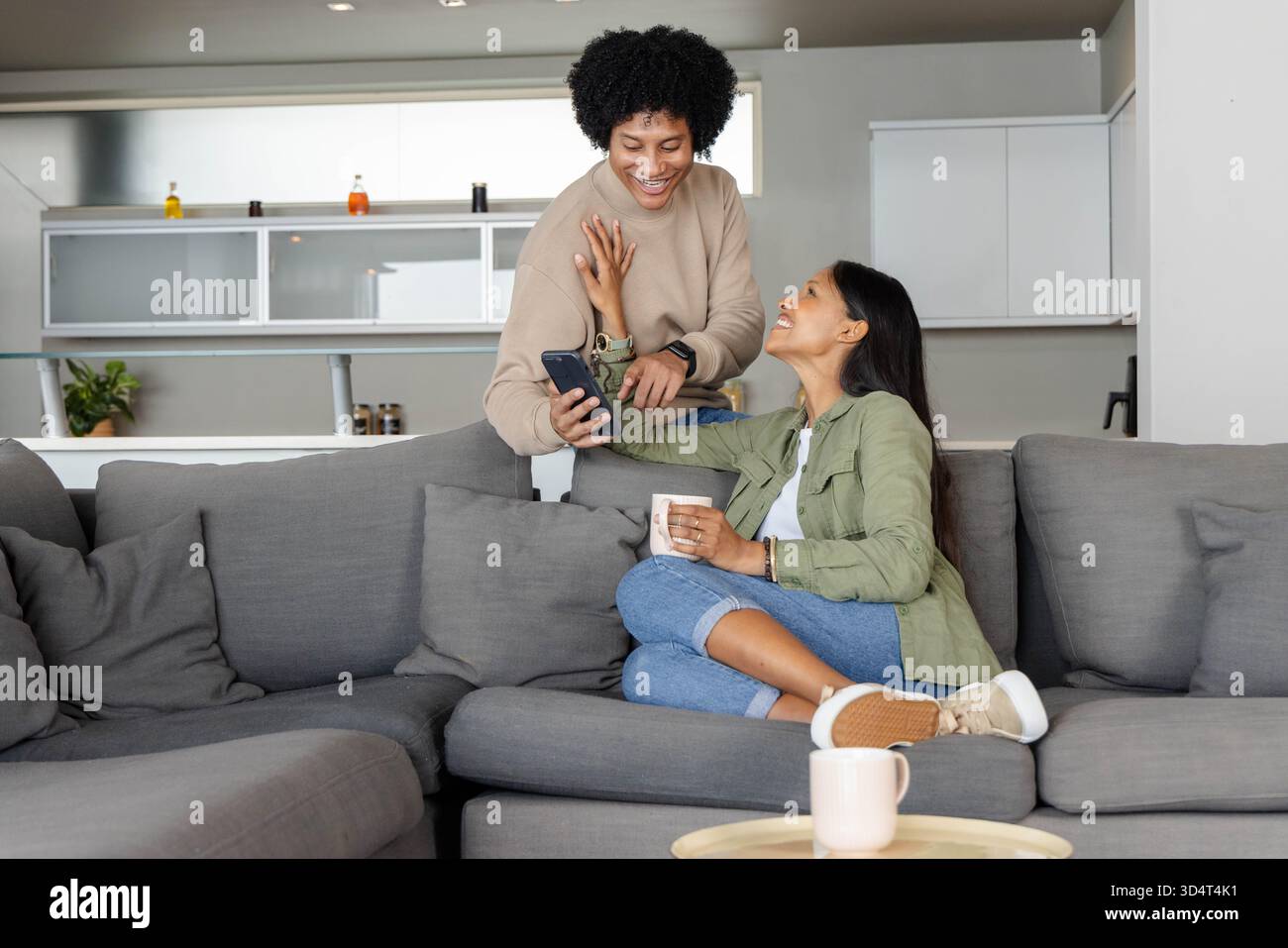Couple diversifié partageant l'écran de smartphone sur le canapé dans le salon ouvert, avec tasses à café Banque D'Images