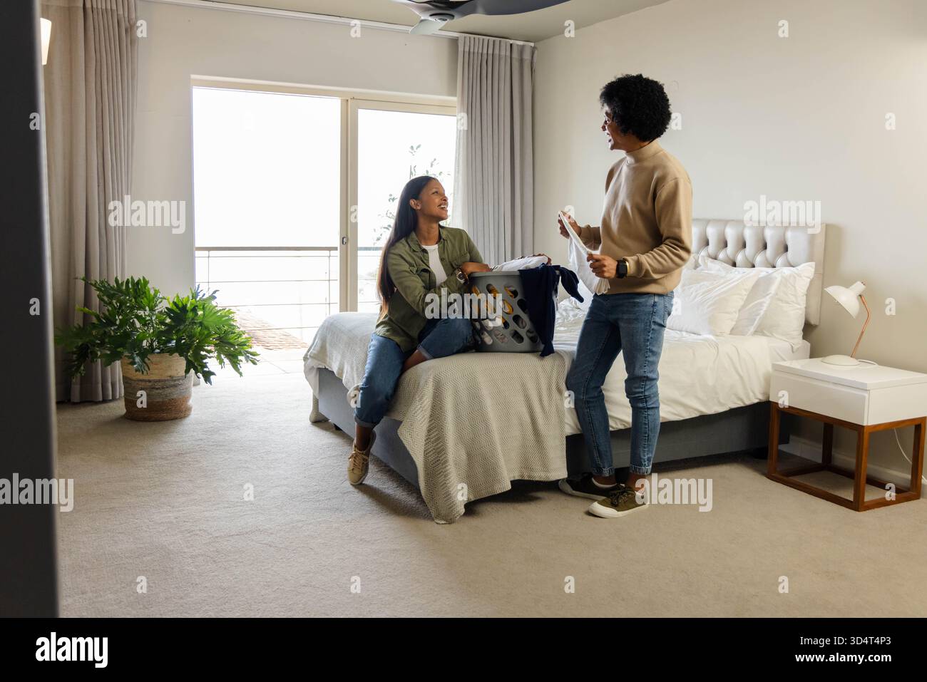 Couple afro-américain triant et pliant le linge sur le lit, avec panier à linge et plante en pot Banque D'Images
