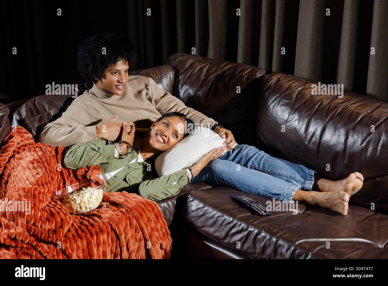 Relaxant couple diversifié se prélasser sur un canapé en cuir dans le salon, avec pop-corn et couverture de jet Banque D'Images