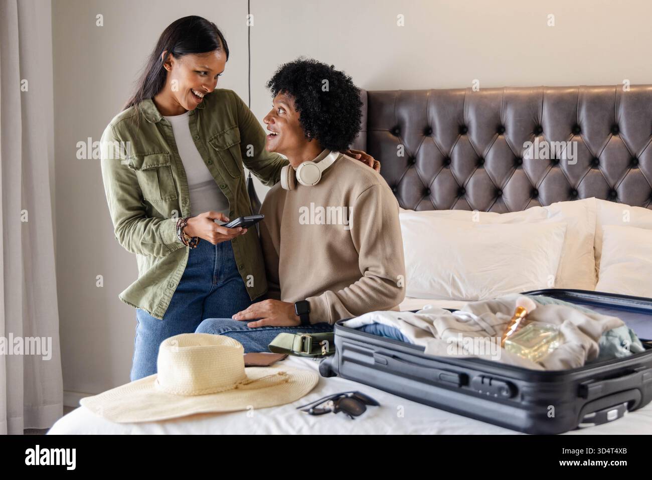 Souriant couple diversifié emballant valise noire sur le lit dans la chambre d'hôtel, avec smartphone et écouteurs Banque D'Images