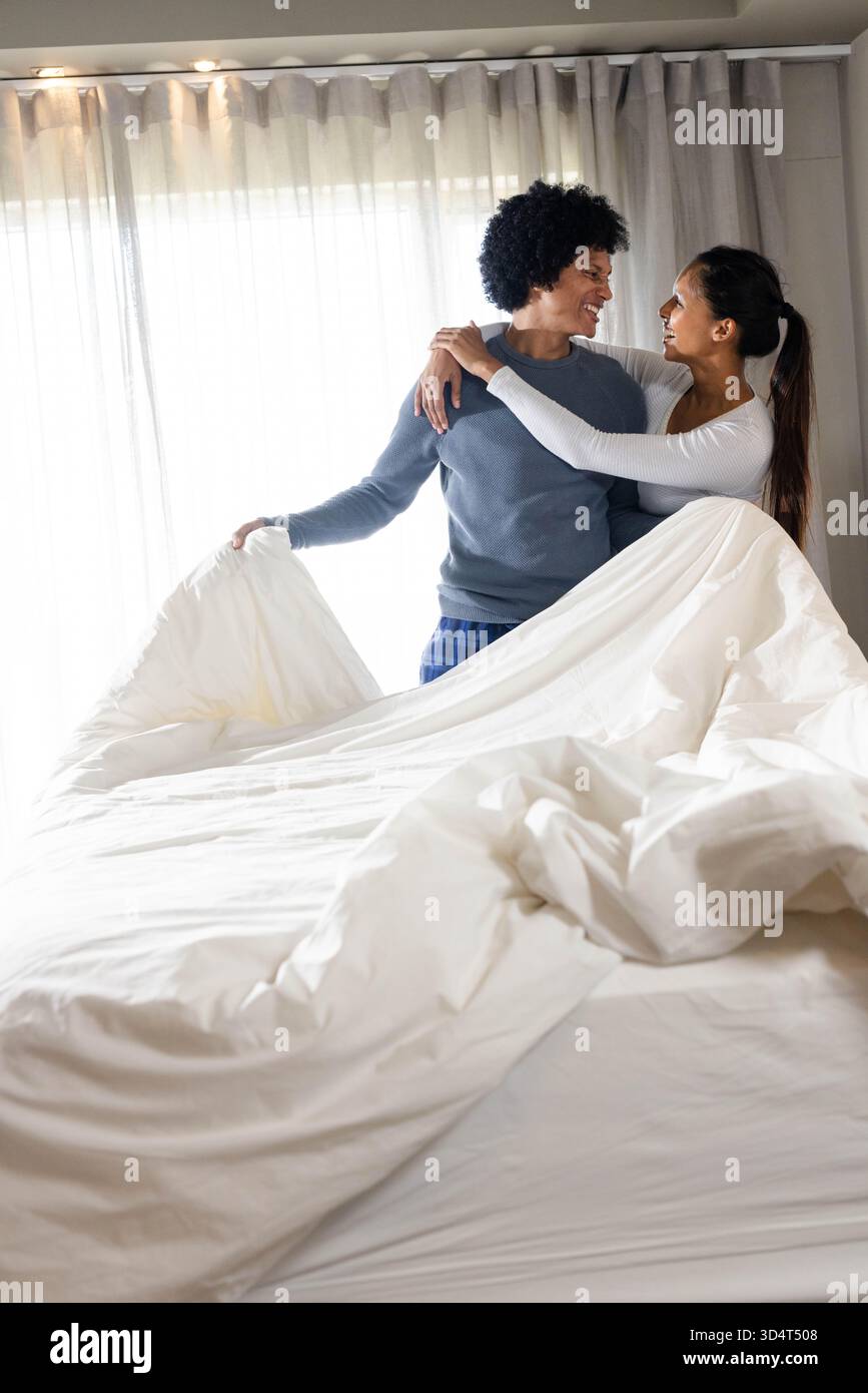 Couple diversifié faisant lit dans une chambre aérée, avec housse de couette blanche et rideaux transparents par fenêtre Banque D'Images