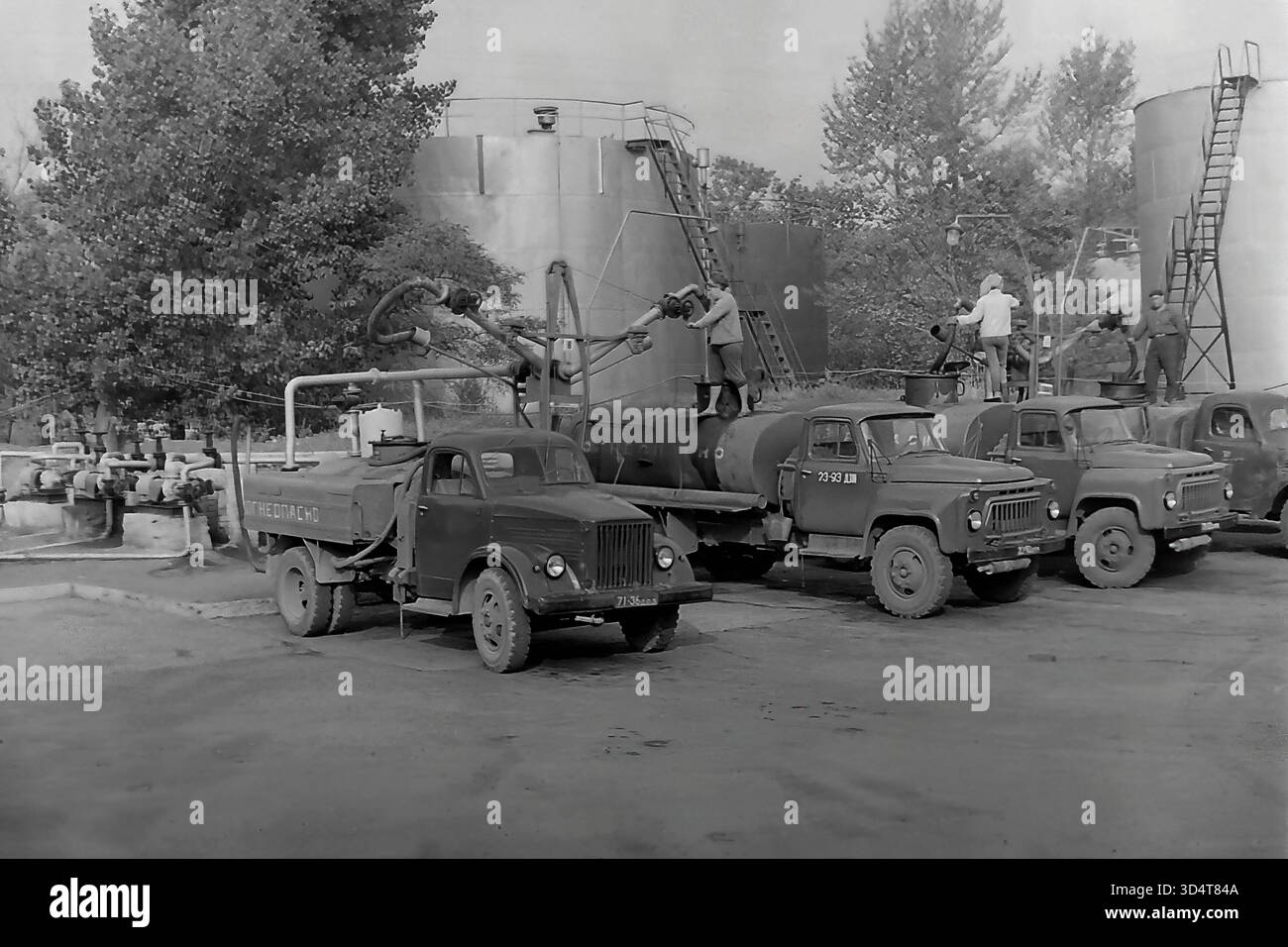Une photo d'archives dynamique en noir et blanc des années 1970 montre la station de chargement animée du dépôt pétrolier de Sloviansk, en RSS d'Ukraine. Une ligne de camions-citernes soviétiques, y compris les modèles gaz-51, est stationnée à côté de grands réservoirs de stockage. Les travailleurs sont activement engagés dans le processus de ravitaillement, debout sur le dessus des citernes et connectant les tuyaux des bras de chargement suspendus. La scène est pleine d'activité et capture les opérations logistiques essentielles du Donbass pacifique. Banque D'Images