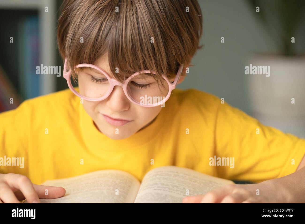 Élève souriant dans des lunettes repose la main tout en lisant un livre ouvert au bureau de l'école. Apprentissage de l'enfance, esprit intelligent, concept d'étude, développement précoce et préparation à l'éducation et à la croissance future. Banque D'Images