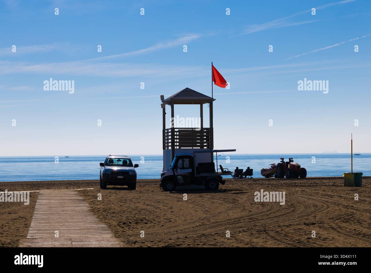 Tour de sauveteur avec drapeau d'avertissement rouge et véhicules de service de plage sur un rivage ensoleillé calme Banque D'Images