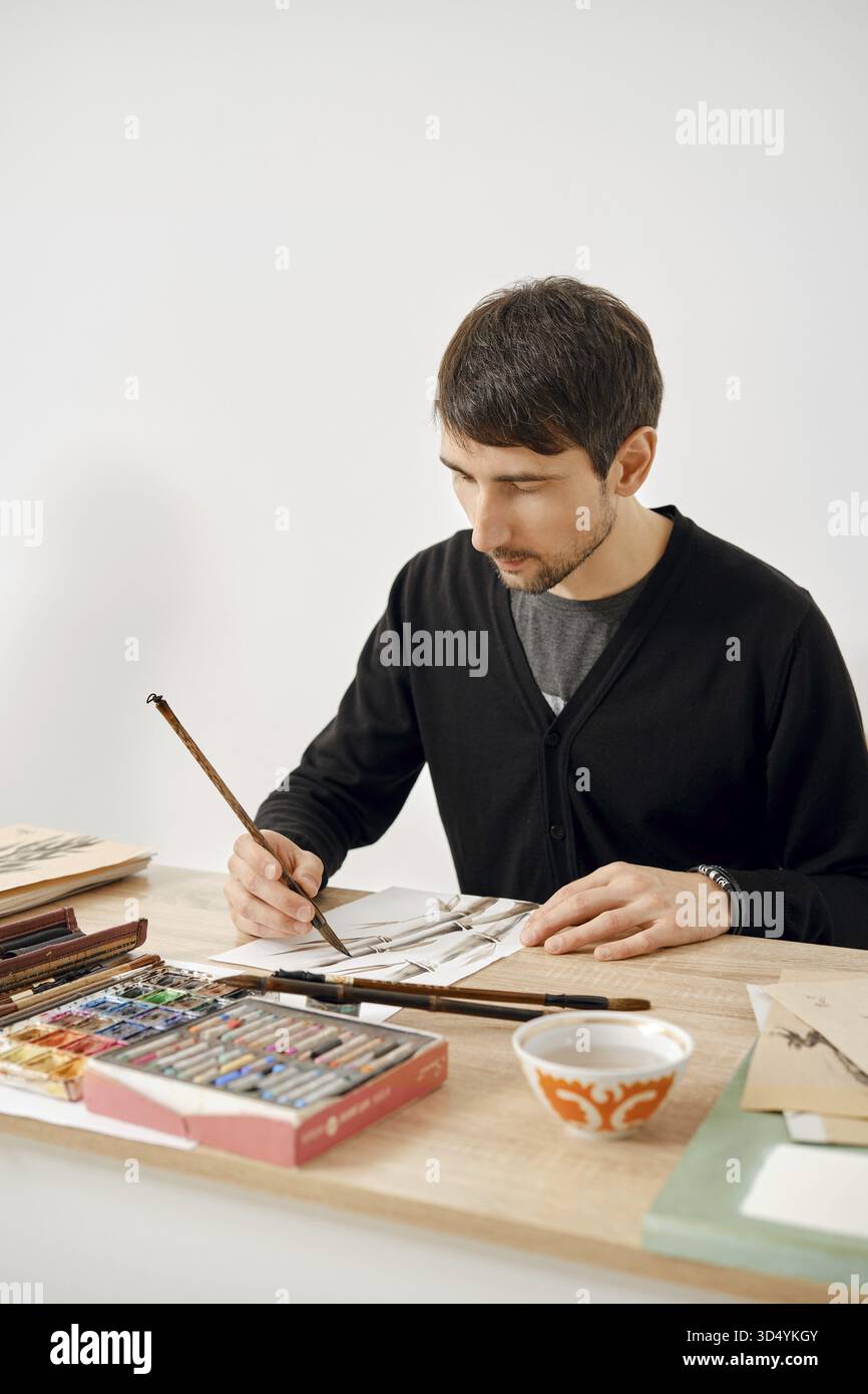 Un artiste masculin se concentre sur son travail à une table en bois remplie de diverses fournitures artistiques. Il utilise des pinceaux et des matériaux colorés pour créer un complexe Banque D'Images