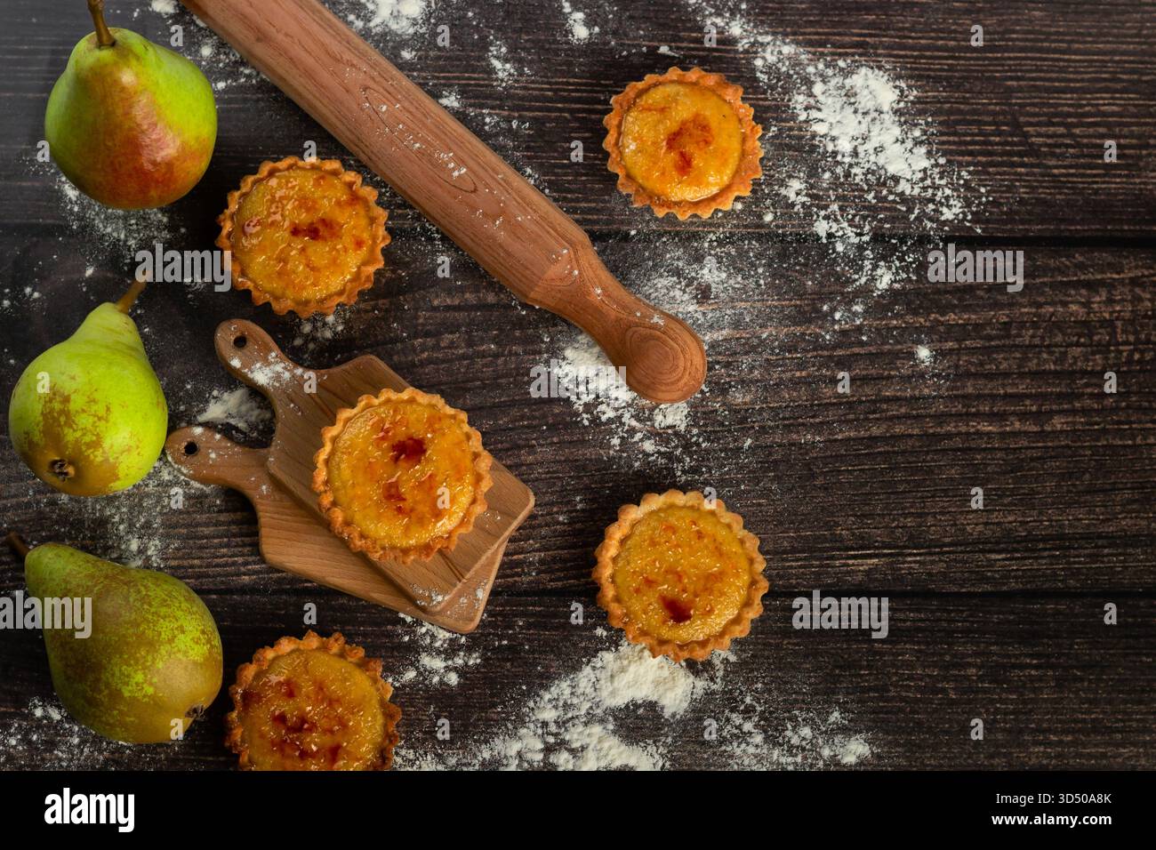 Les tartes aux poires fraîchement cuites reposent sur une planche à découper en bois, entourées de poires et de farine. Un rouleau à pâtisserie repose à proximité, créant une atmosphère chaleureuse et accueillante Banque D'Images