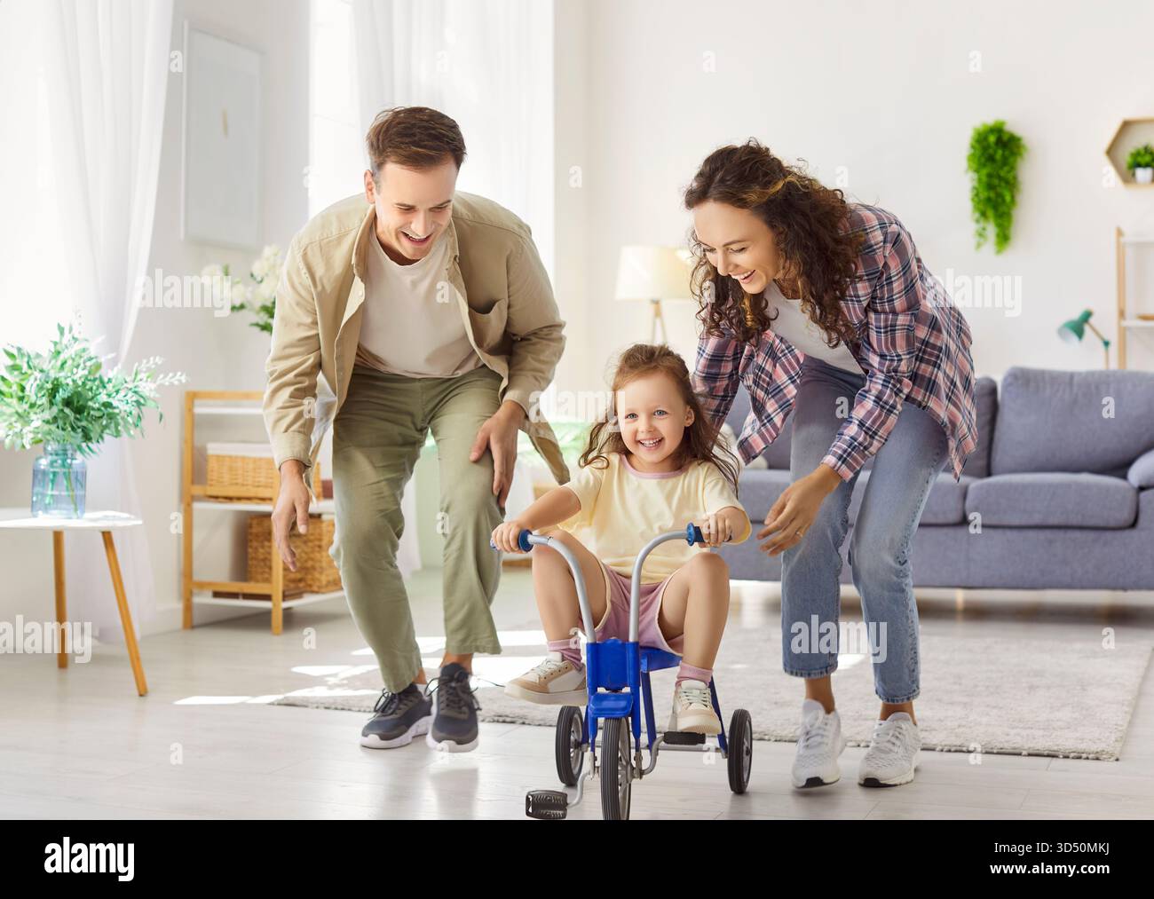 Les parents heureux aident leur fille à monter tricycle à l'intérieur, moment familial ludique à la maison Banque D'Images
