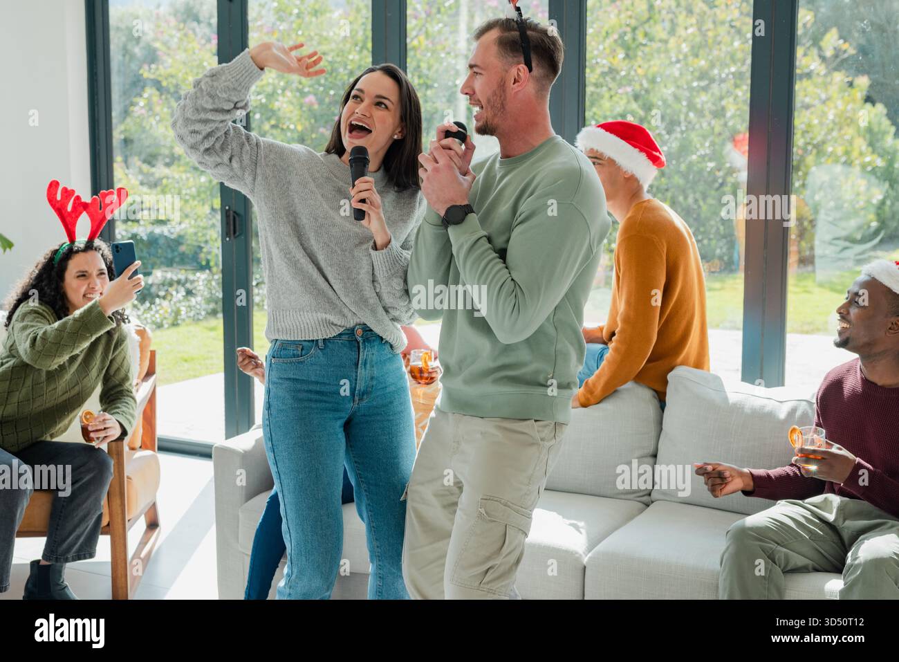 Divers amis chantant karaoké dans le salon à la maison, portant des chapeaux festifs, tenant des microphones Banque D'Images