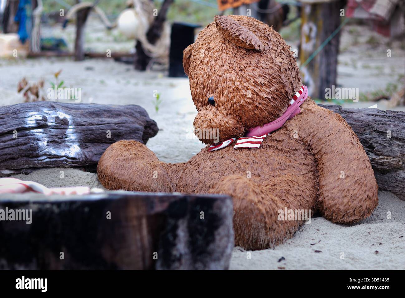 Un grand ours en peluche brun saturé avec une écharpe rayée rose et blanche est assis avec découragement ou contemplation dans le sable humide sur une plage Banque D'Images