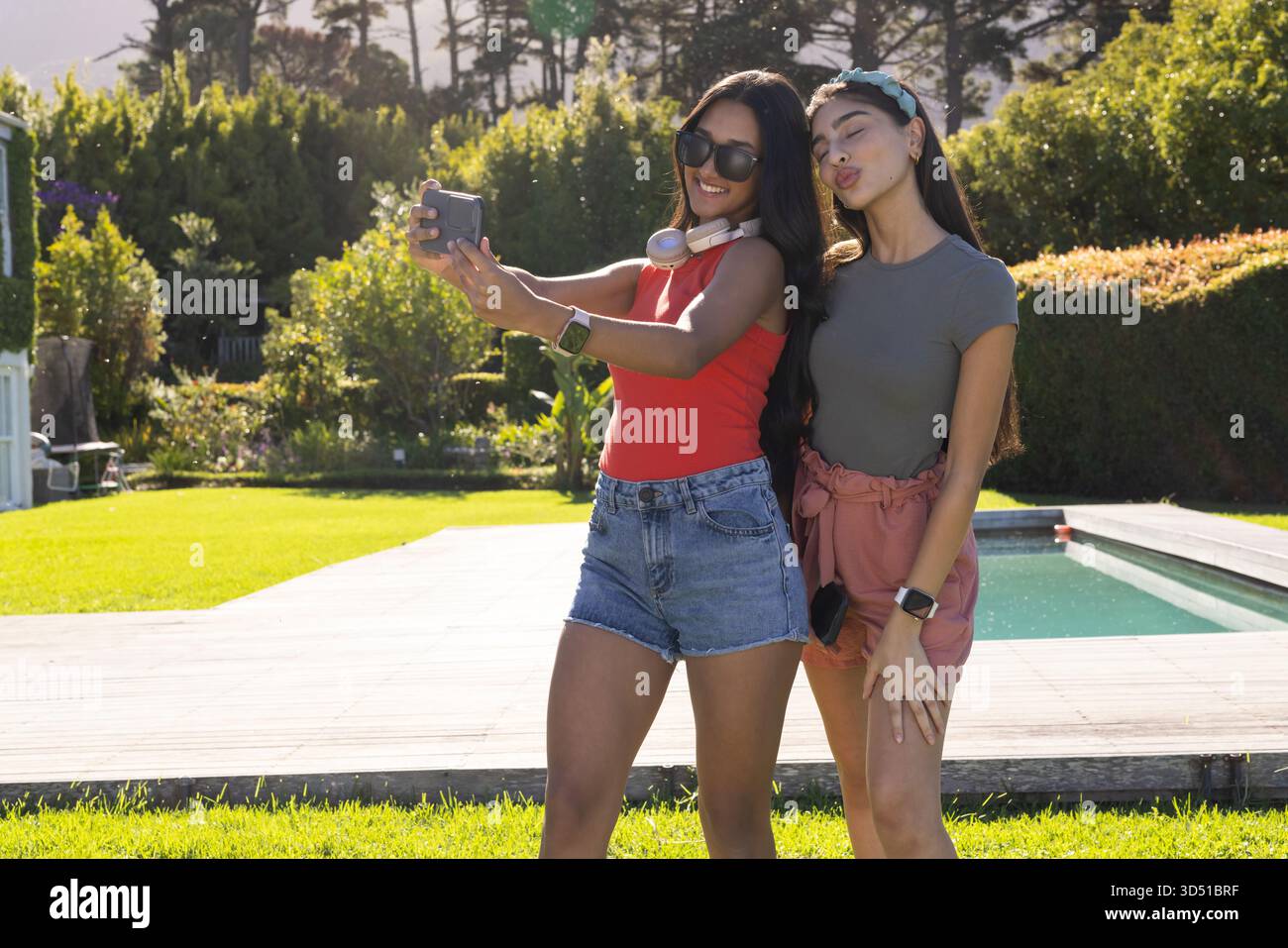 Selfie diverses amies féminines debout près de la terrasse de la piscine arrière, avec smartphone et écouteurs Banque D'Images