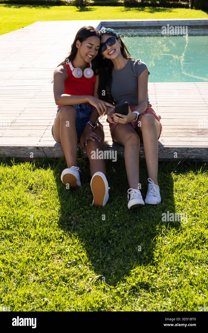 Assis diverses amies se penchant sur la terrasse en bois au bord de la piscine, à l'aide d'un smartphone et d'un casque Banque D'Images