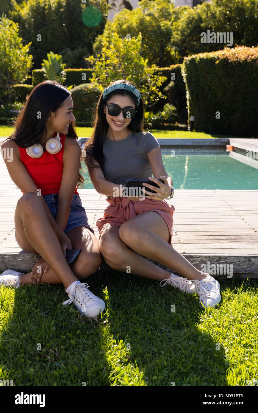 Souriant diverses amies se penchant vers un smartphone au bord de la piscine, avec des écouteurs, des lunettes de soleil Banque D'Images