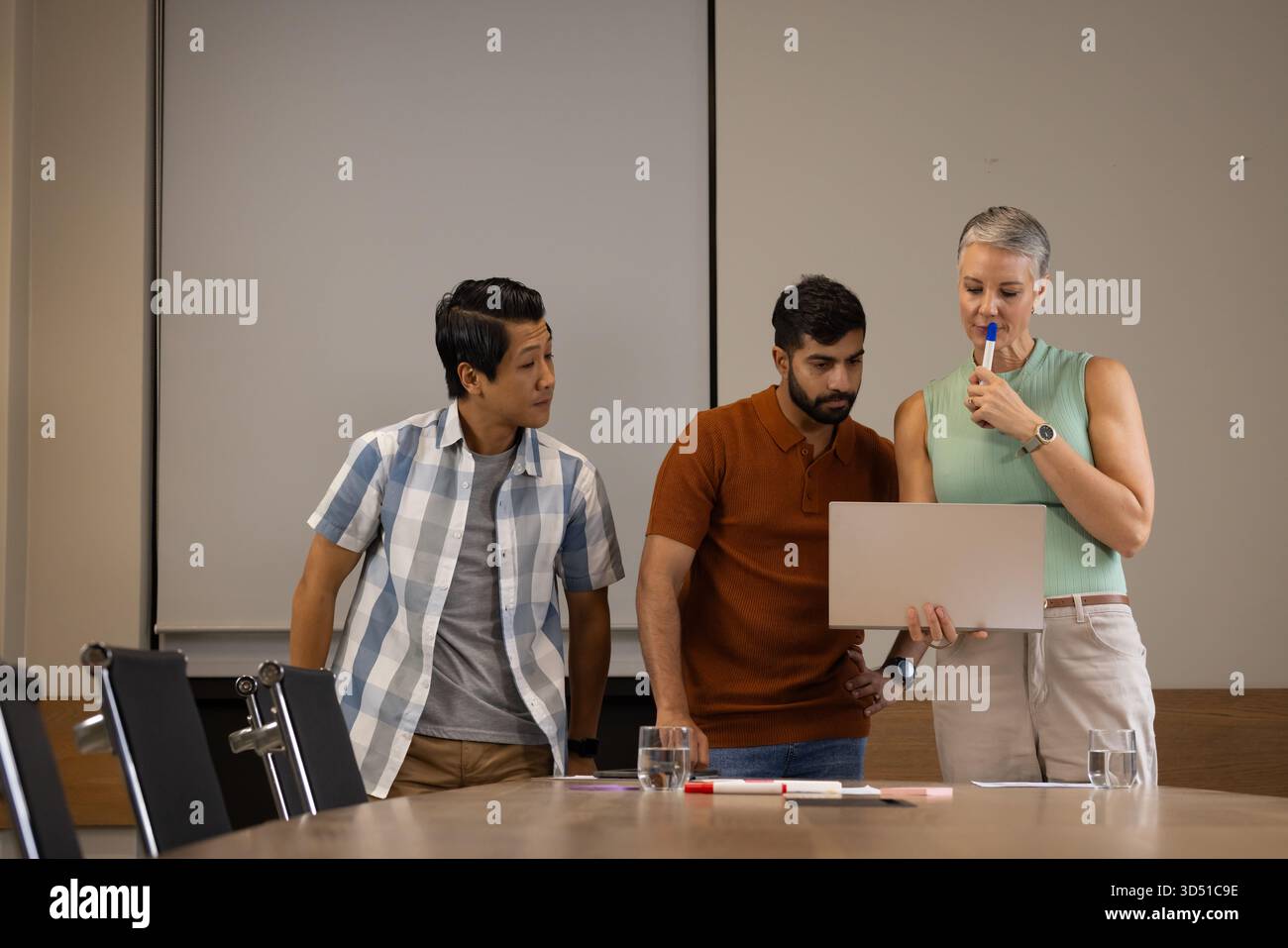 Examen de divers collègues penchés sur l'ordinateur portable dans la salle de réunion, avec des marqueurs et des documents colorés Banque D'Images