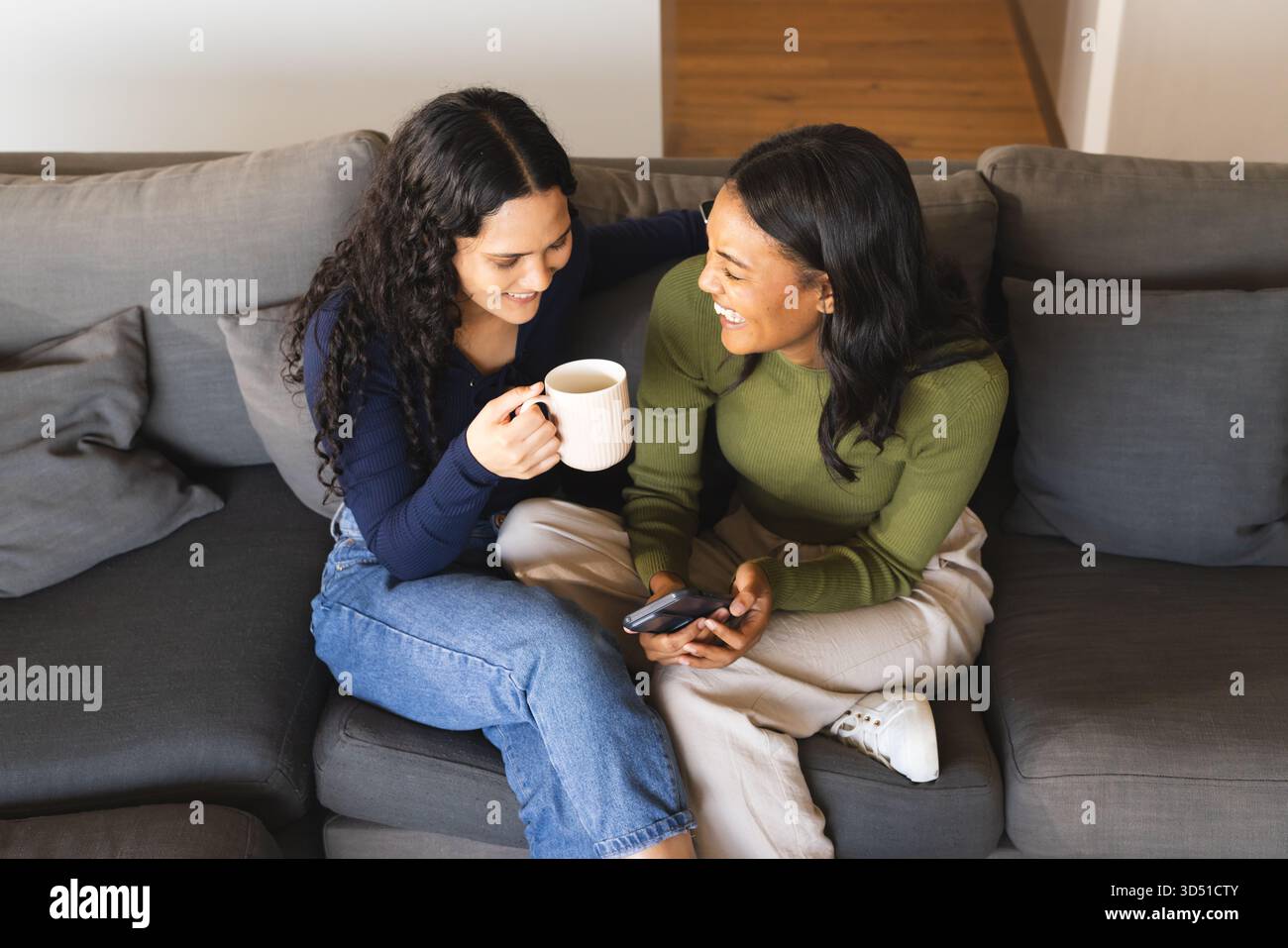 Rire diverses amies féminines assises sur un canapé gris dans le salon, tenant une tasse et un smartphone Banque D'Images