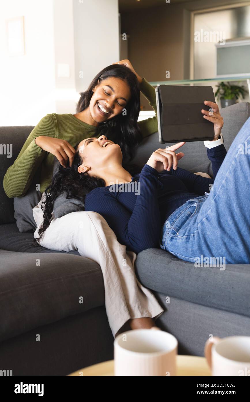 Riant diverses amies féminines pointant vers la tablette sur le canapé gris, avec une table basse et des tasses blanches Banque D'Images