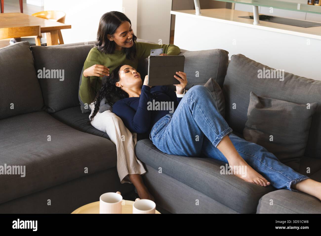 Relaxer divers partenaires féminins partageant tablette sur canapé dans le salon, avec smartphone et tasses Banque D'Images