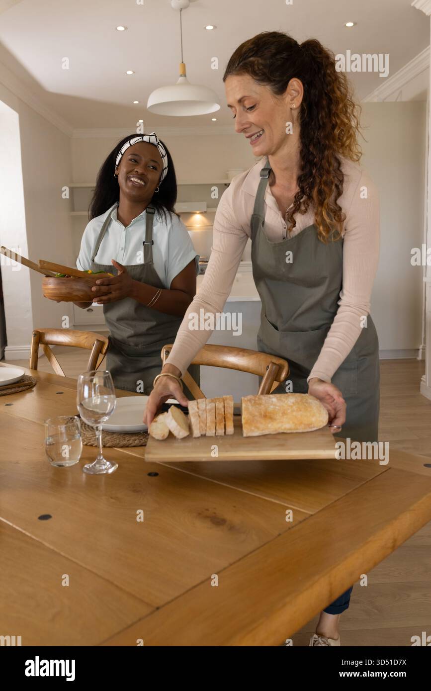 Diverses amies féminines arrangeant un bol à salade et du pain sur une planche à découper dans la cuisine à la maison, avec des assiettes Banque D'Images
