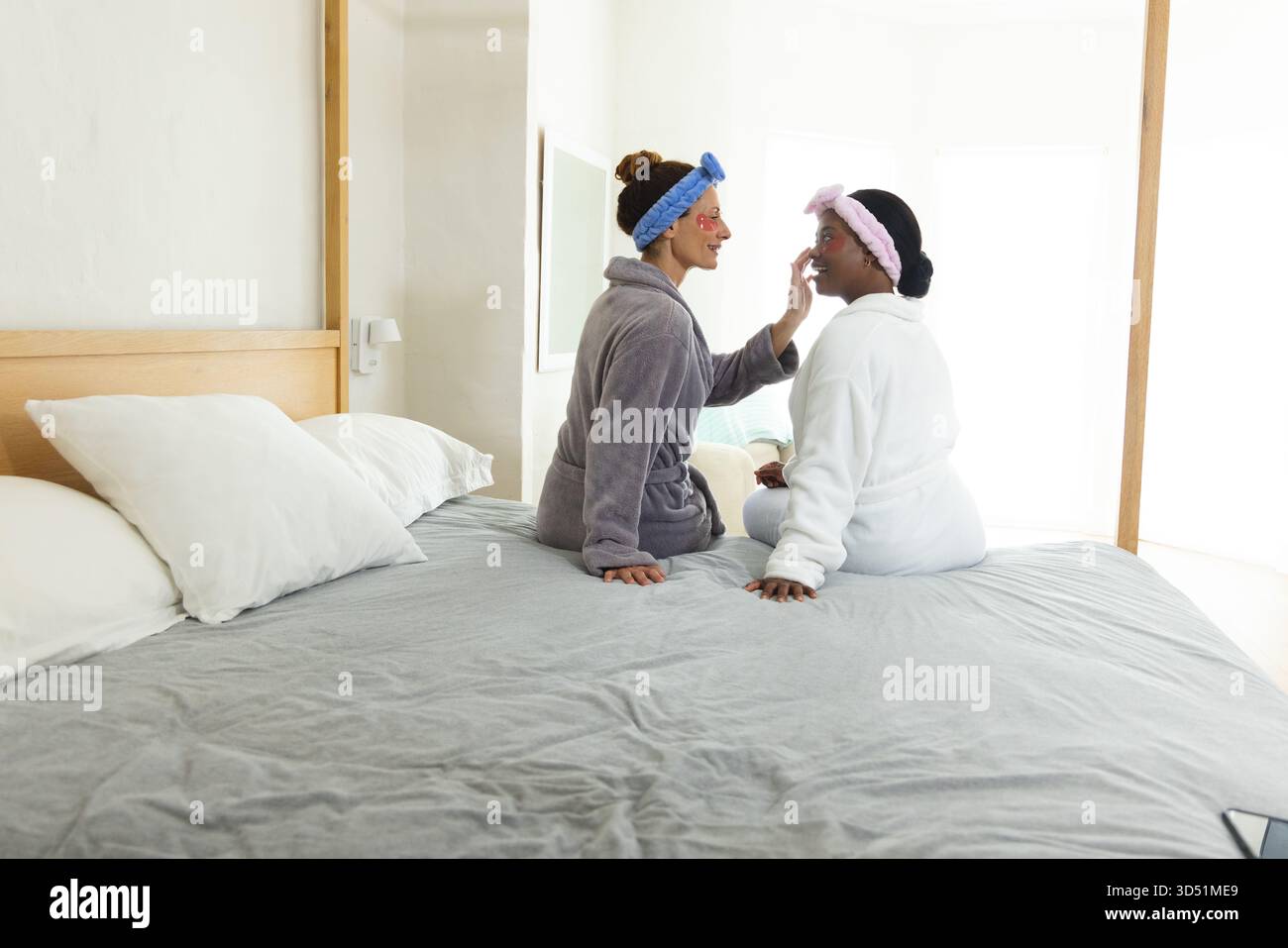 Diverses amies féminines appliquant des masques faciaux rouges sur le lit dans une chambre lumineuse, avec bandeaux Banque D'Images
