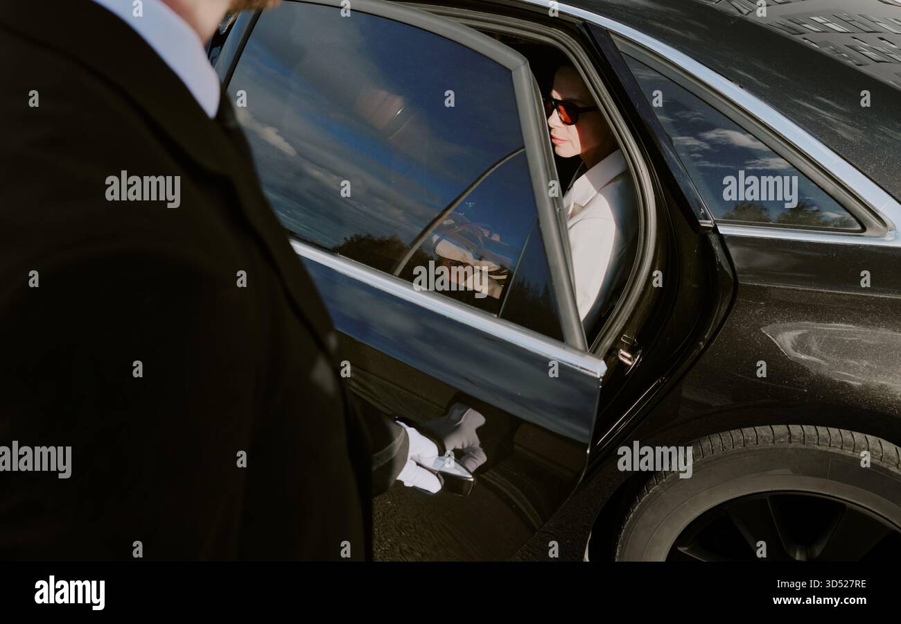 Jeune homme adulte portant des lunettes de soleil sortant de la voiture assisté par chauffeur Banque D'Images