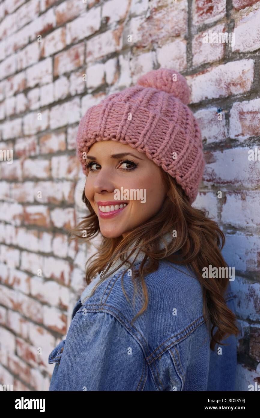 Portrait d'une femme souriante portant une veste en denim et un chapeau en laine debout devant un mur de briques Banque D'Images