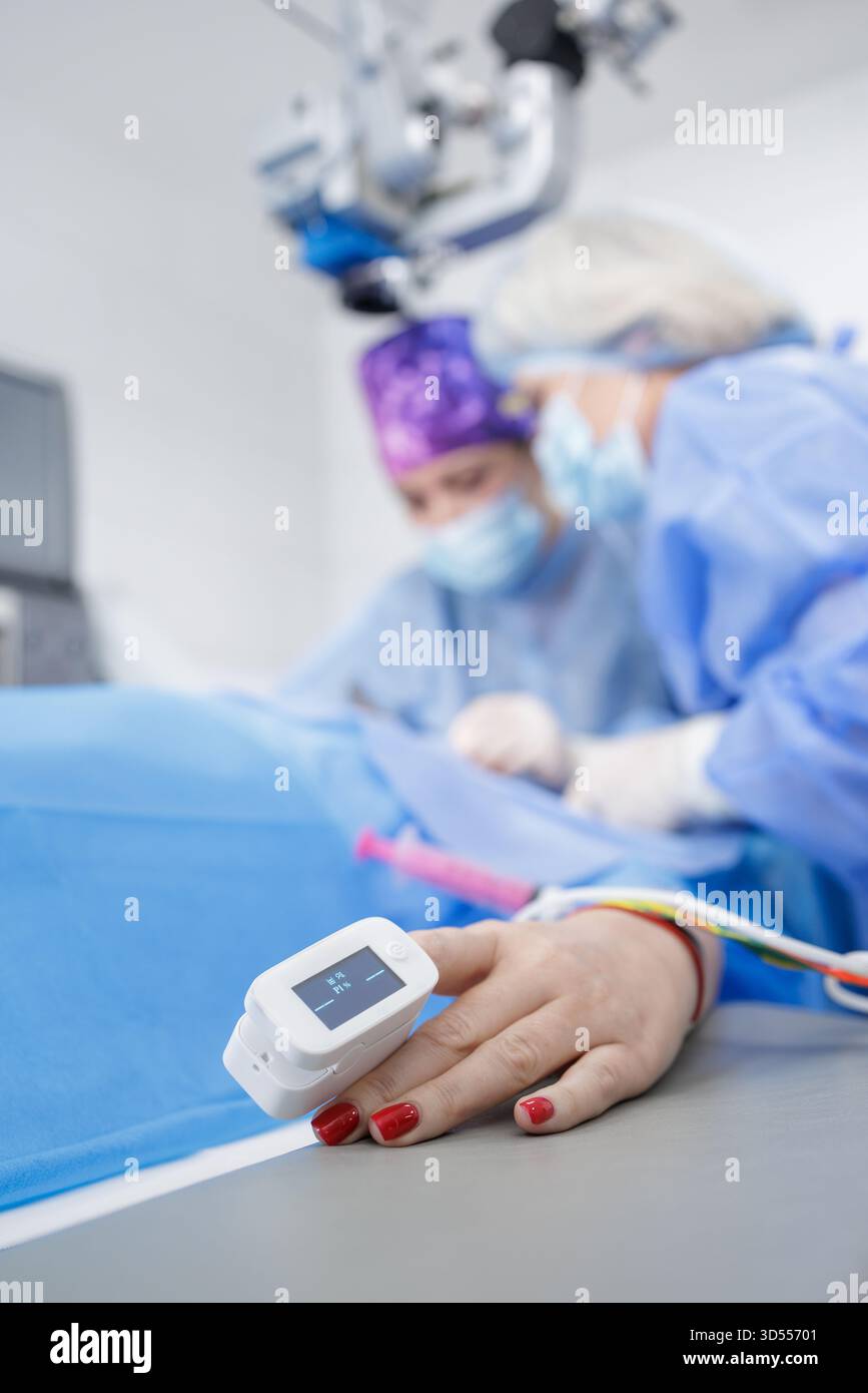 Gros plan sur la main avec oxymètre de pouls pendant que l'équipe effectue la chirurgie des paupières, montrant la surveillance périopératoire et la sécurité dans une salle d'opération moderne. Banque D'Images