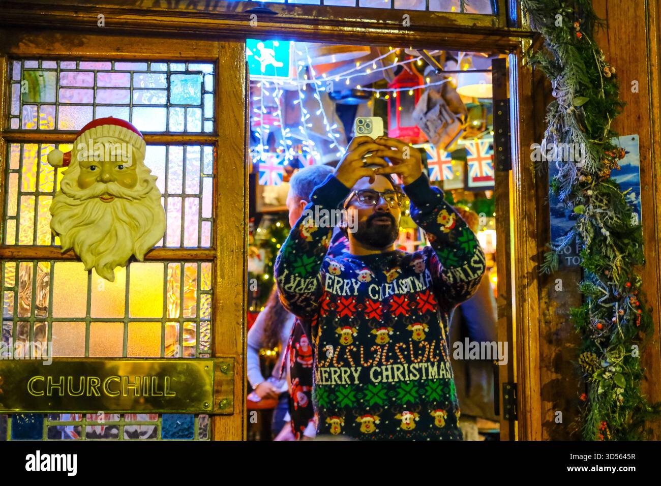 Londres, Royaume-Uni. 13 novembre 2025. Un membre du personnel prend des photos de la foule à l'extérieur. Les Churchill Arms de Kensington mettent en marche leur exposition festive annuelle très appréciée, composée de plus de 80 sapins et de milliers de lumières, ce qui leur a valu le titre de « pub le plus chrétien » de Londres. La pluie n'a pas assombri les esprits de la foule, et le directeur général James Keogh a soigneusement vérifié si des bus passaient avant le compte à rebours, après qu'une vidéo virale humoristique filmée l'année dernière sur la route en a montré un bloquant la vue d'un spectateur au moment exact où les lumières se sont allumées. Crédit : onzième heure photographie/Alamy Banque D'Images