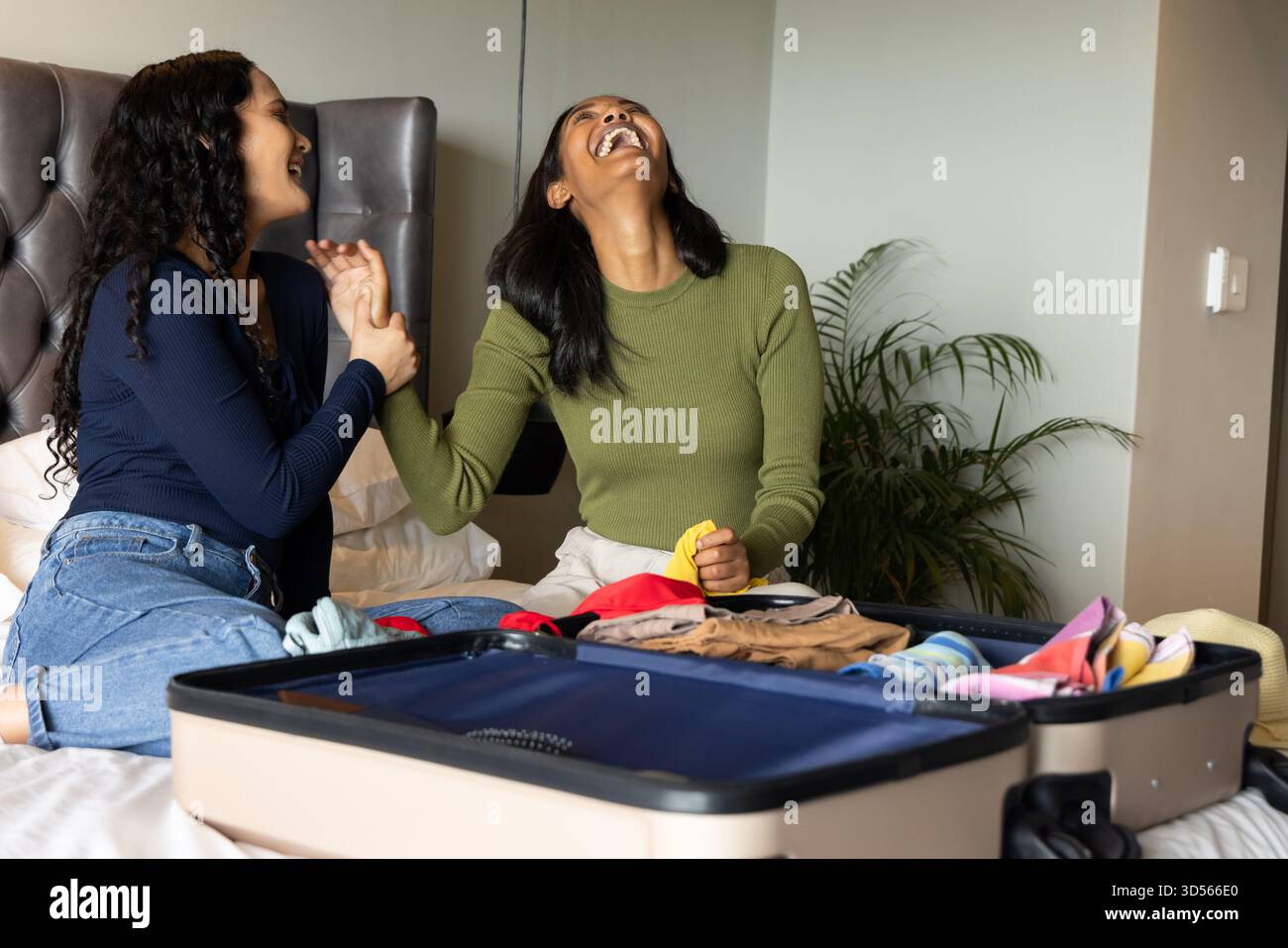 Rire diverses amies féminines emballant des vêtements sur le lit dans la chambre à coucher, avec valise ouverte et plante Banque D'Images