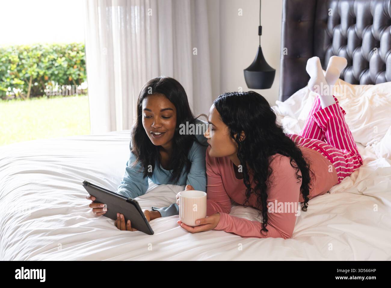 Relaxant diverses amies féminines allongées sur le lit dans une chambre lumineuse, avec tablette et tasse blanche Banque D'Images