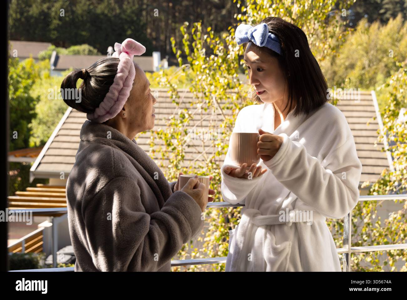 Mère aînée asiatique et fille dans des peignoirs appréciant le café sur le balcon, souriant chaleureusement Banque D'Images