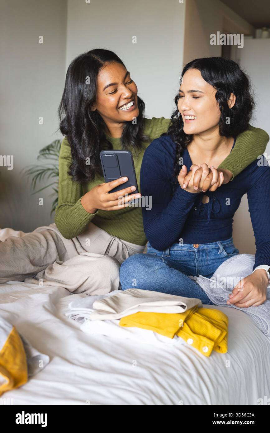 Blotti diverses amies féminines souriant sur le lit dans une chambre confortable, avec smartphone et vêtements pliés Banque D'Images