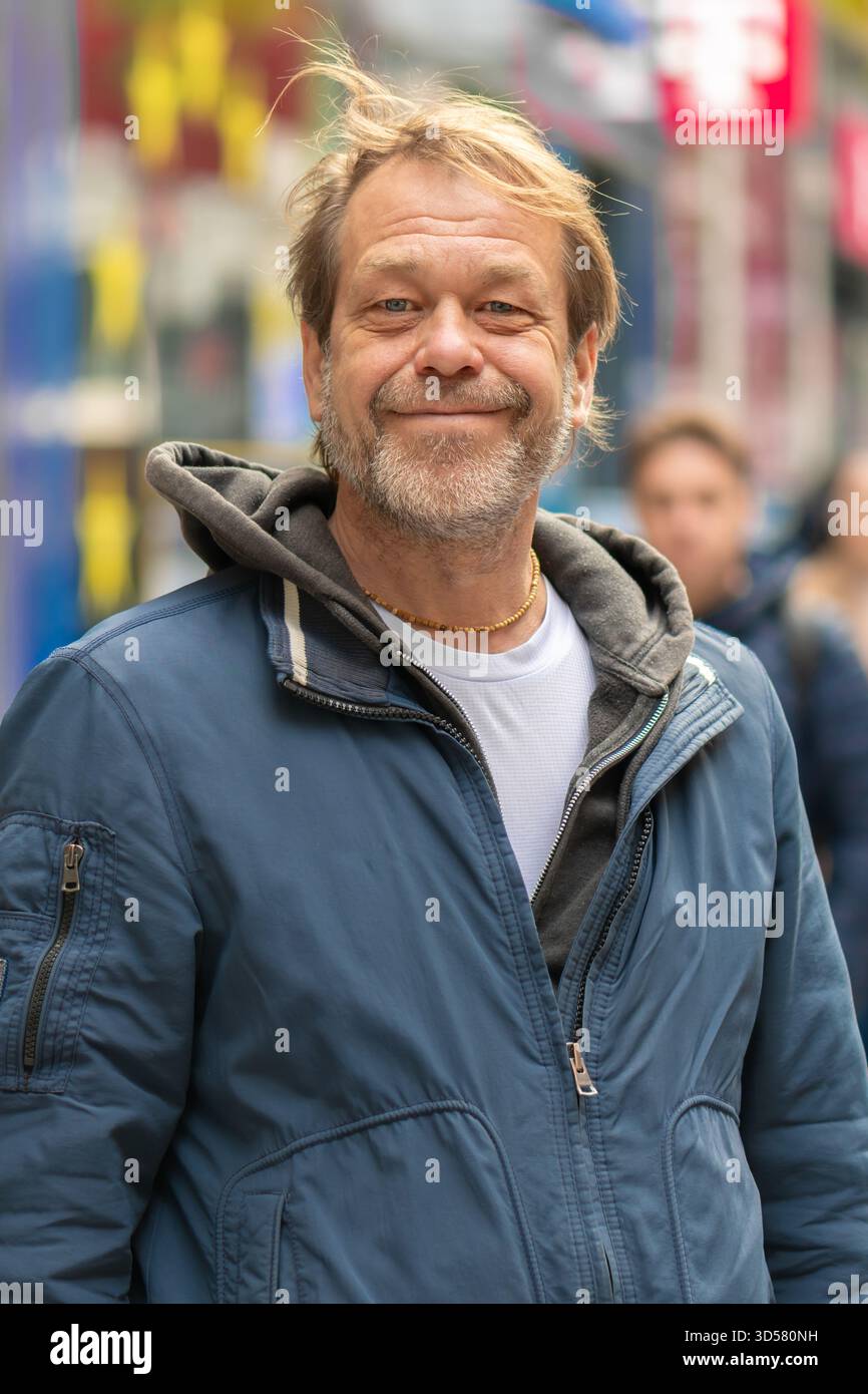 Un homme de 50 à 55 ans en veste sourit alors qu'il se tient debout dans une rue animée de la ville parmi les passants Banque D'Images