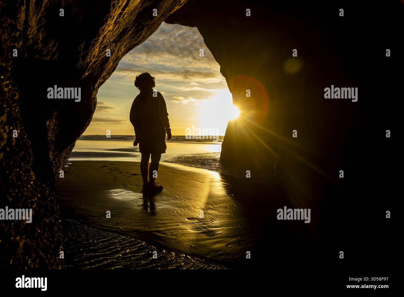 Vue d'un personnage silhouetté contre le coucher de soleil radieux vu d'une entrée de grotte sombre, le sable humide reflétant la lumière dorée, Tongaporutu, Taran Banque D'Images