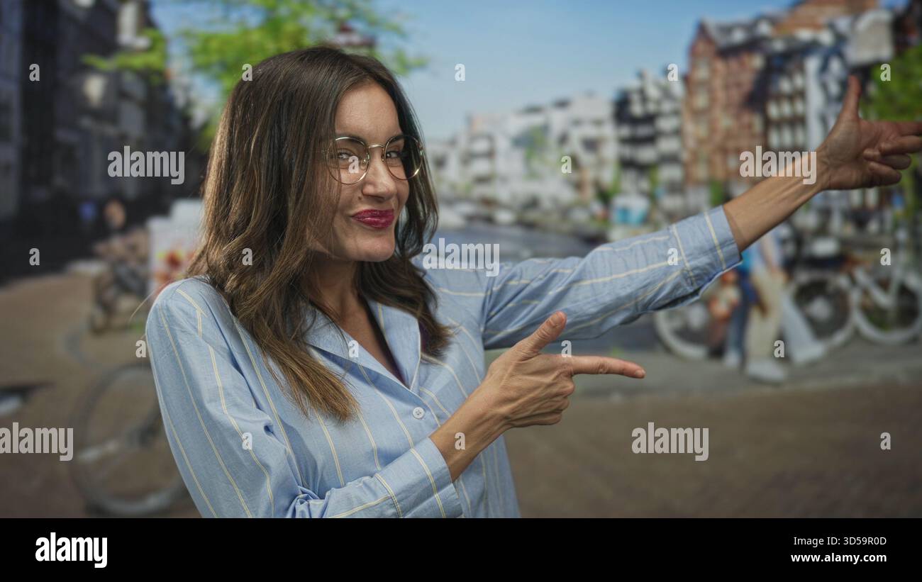 Femme souriante en chemise bleue avec des lunettes et un large sourire du doigt pointant vers le canal ensoleillé sur la rue ; joie insouciante. Banque D'Images
