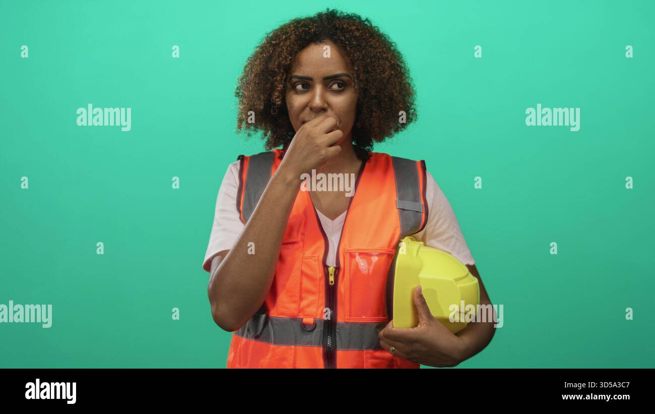 Femme ingénieur tenant un casque jaune avec le doigt sur les lèvres dans le studio sarcelle ; conscience de la sécurité réfléchie. Banque D'Images