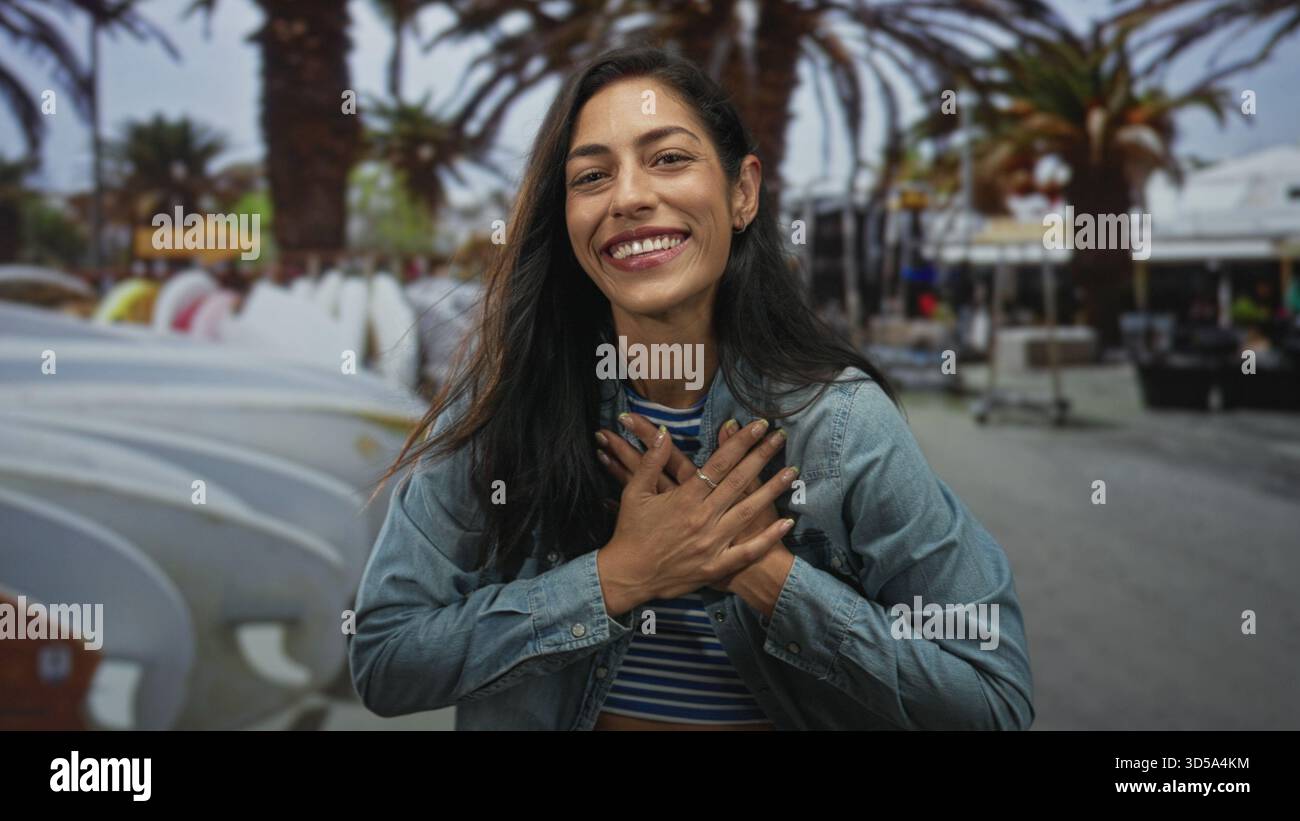 Femme avec les mains croisées sur la poitrine portant une veste en denim et un crop top rayé sur la rue avec des planches de surf et des palmiers ; connexion de gratitude. Banque D'Images