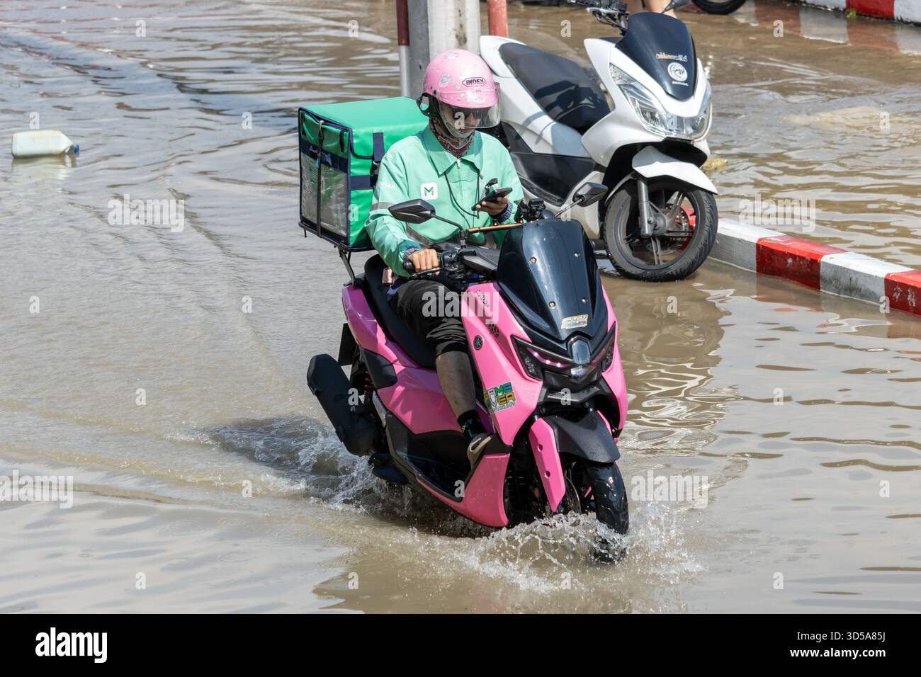 BANGKOK, THAÏLANDE, nov 09 2025, Un livreur sur un scooter rose traverse une rue inondée de la ville à l'aide d'un téléphone portable Banque D'Images