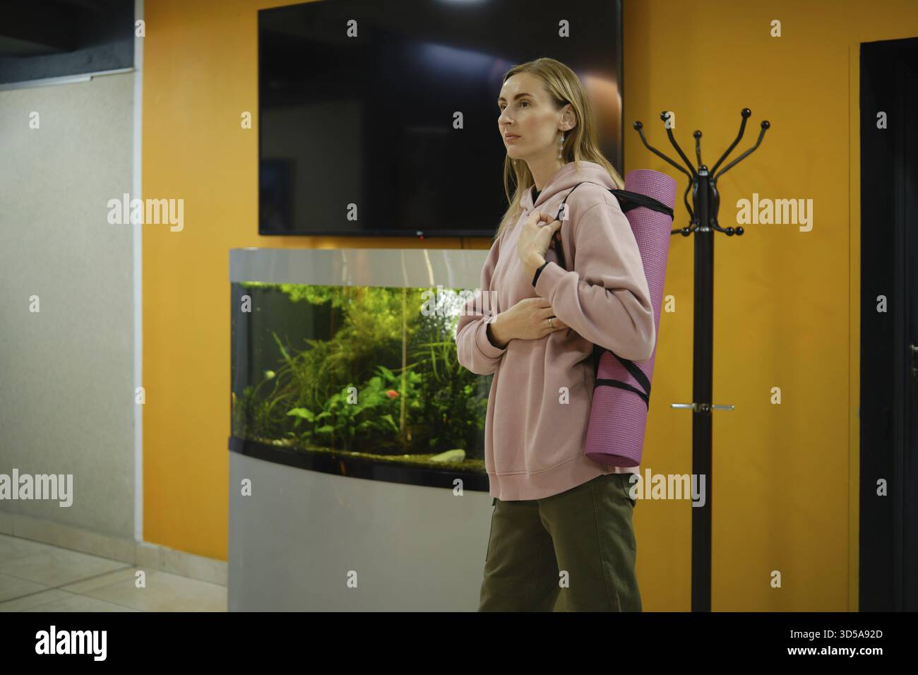 Une femme se tient debout dans un studio de fitness tenant un tapis de yoga. Elle porte un sweat à capuche confortable et semble prête pour son cours de yoga. Un aquarium ajoute de la vie à l'espace Banque D'Images