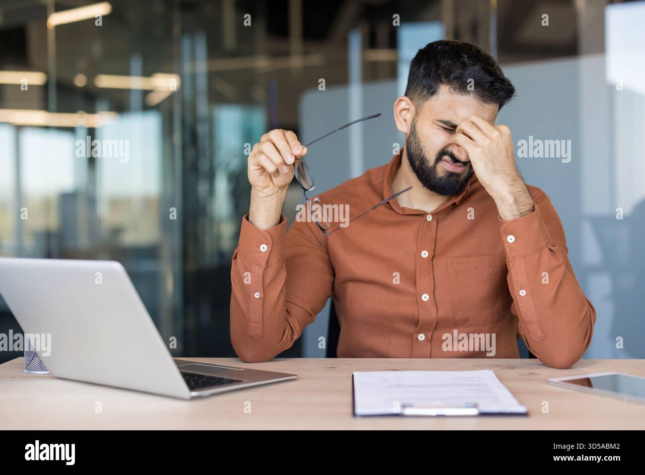 Jeune homme se sentant fatigué et ayant un mal de tête douloureux, frottant ses tempes dans l'inconfort tout en tenant des lunettes, éprouvant l'épuisement professionnel et la fatigue oculaire au bureau Banque D'Images