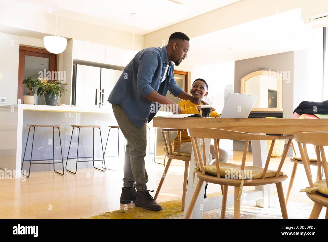 Père afro-américain et fils discutant près de l'ordinateur portable et tasse sur la table à manger dans la cuisine moderne Banque D'Images
