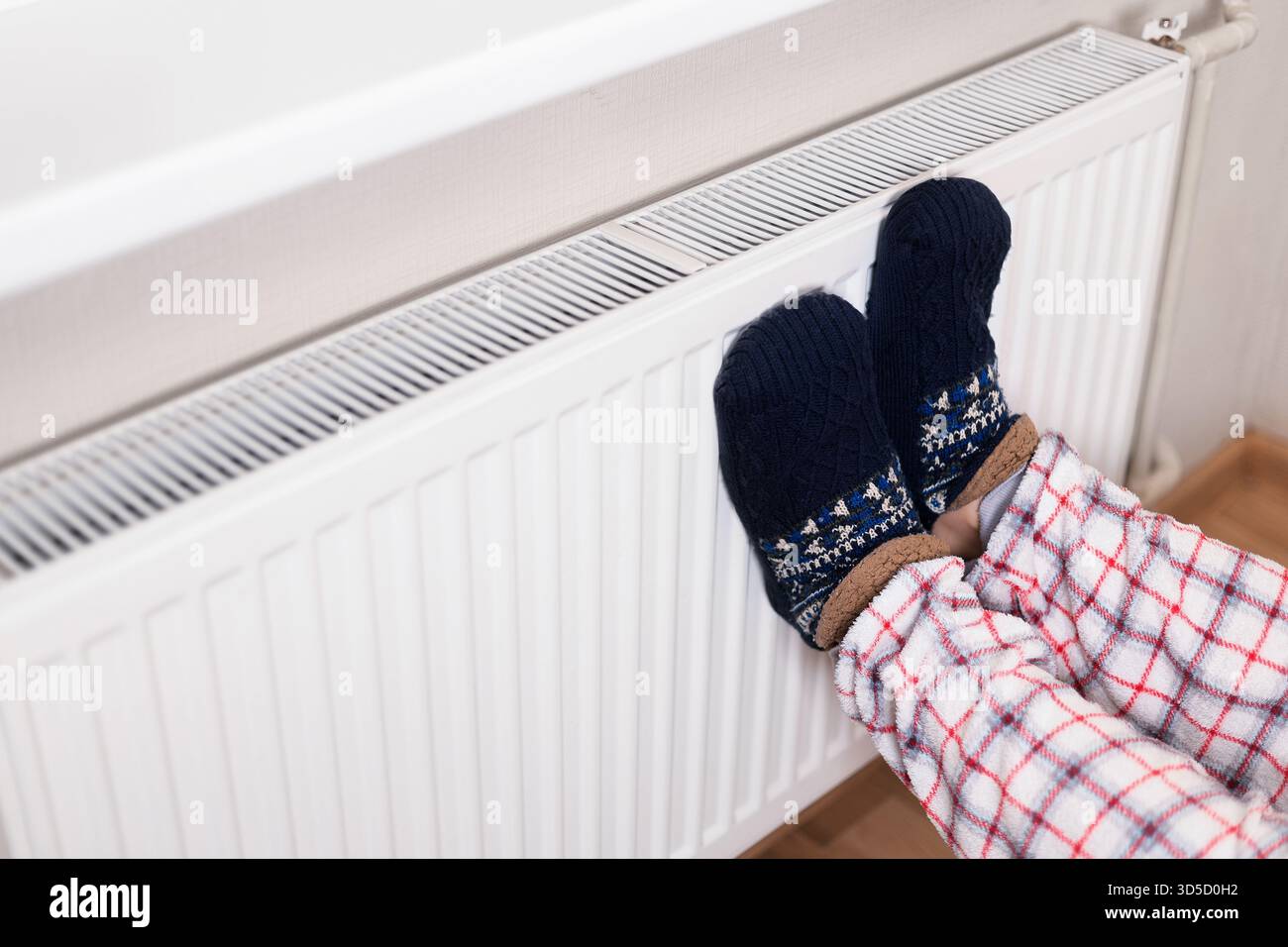 Personne portant des chaussons à motifs confortables et pyjama à carreaux reposant les pieds sur un radiateur blanc pour se réchauffer dans le froid. Photo de haute qualité Banque D'Images